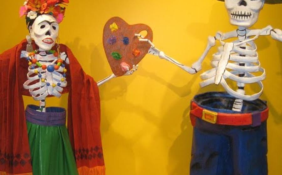 Two skeletons dressed up against a yellow wall. There is text on the wall that says All You Need to Know to Teach Dia de los Muertos, Day of the Dead